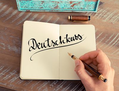 Ein aufgeschlagenes Notizbuch liegt auf einem Tisch. Eine Hand hält einen Füller. In Schreibschrift ist das Wort "Deutschkurs" über beide Seiten in das Notizbuch geschrieben.