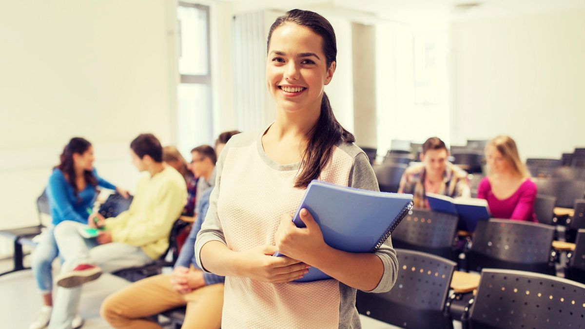 Eine junge Studentin steht lächelnd frontal vor der Kamera. Sie hält einen Schreibblock im Arm. Im Hintergrund sind weitere Studierende und leere Stühle zu sehen.