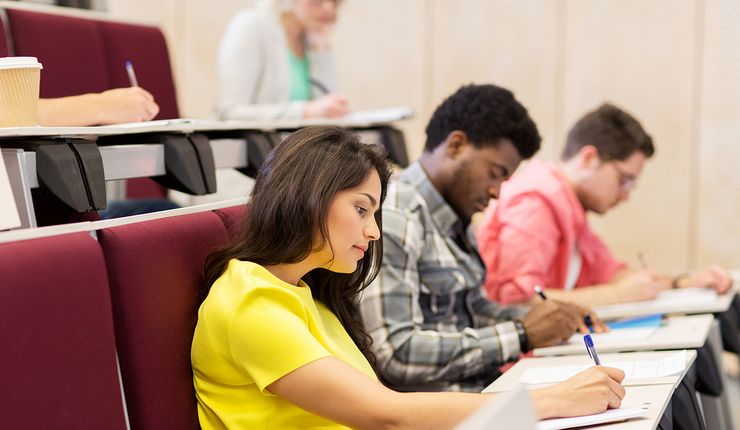 Eine Sitzreihe im Hörsaal, an den Klapptischen sitzen eine junge Frau und daneben zwei junge Männer und schreiben etwas auf.