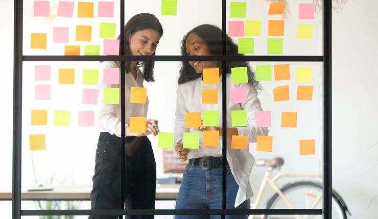 Zwei Junge Frauen stehen vor einer Glaswand, an der Post-its kleben, und besprechen die Notizen vor ihnen.