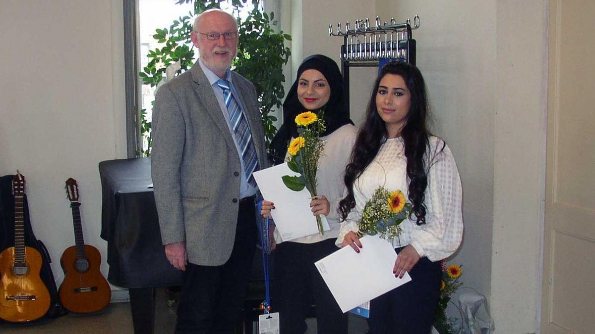 Ein älterer Mann im Anzug gratuliert zwei jungen syrischen Frauen, die ihr Zeugnis in der Hand halten.